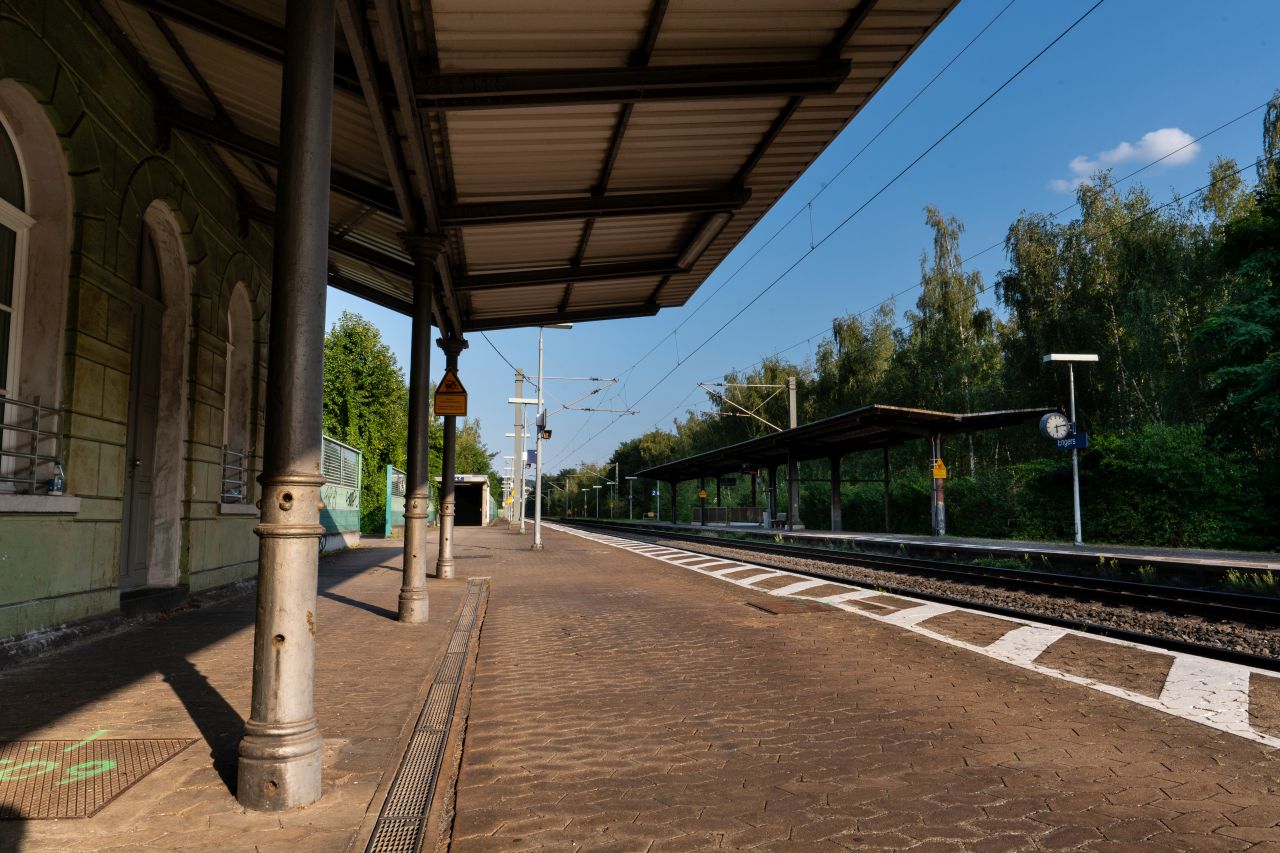 Bahnsteig des Bahnhof Engers mit einem überdachten Wartebereich, Bäumen und Gleisen an einem sonnigen Tag.