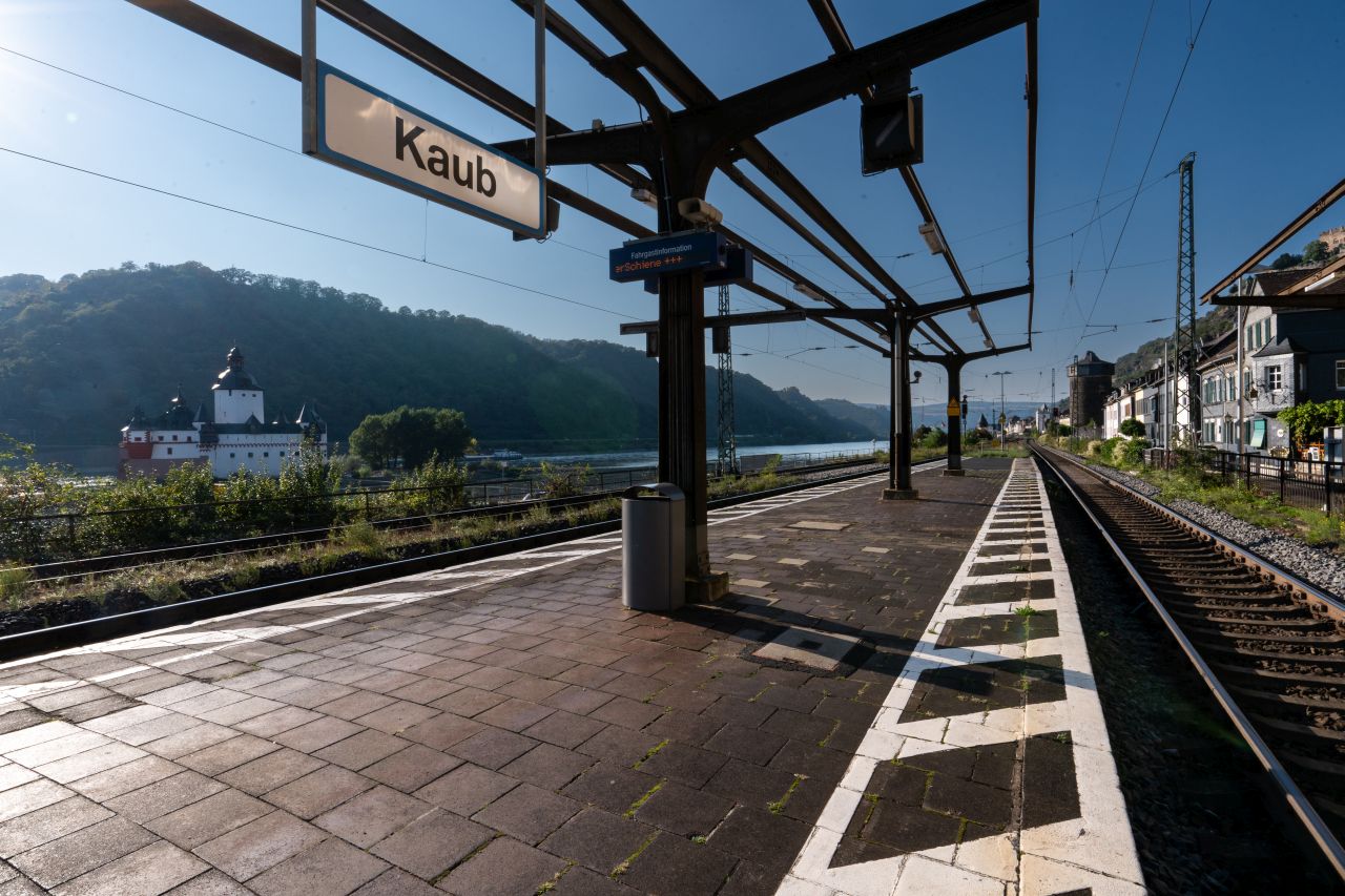 Leerer Bahnsteig mit einem Schild, auf dem "Kaub" steht, und Gleisen neben einem Fluss und grünen Hügeln.