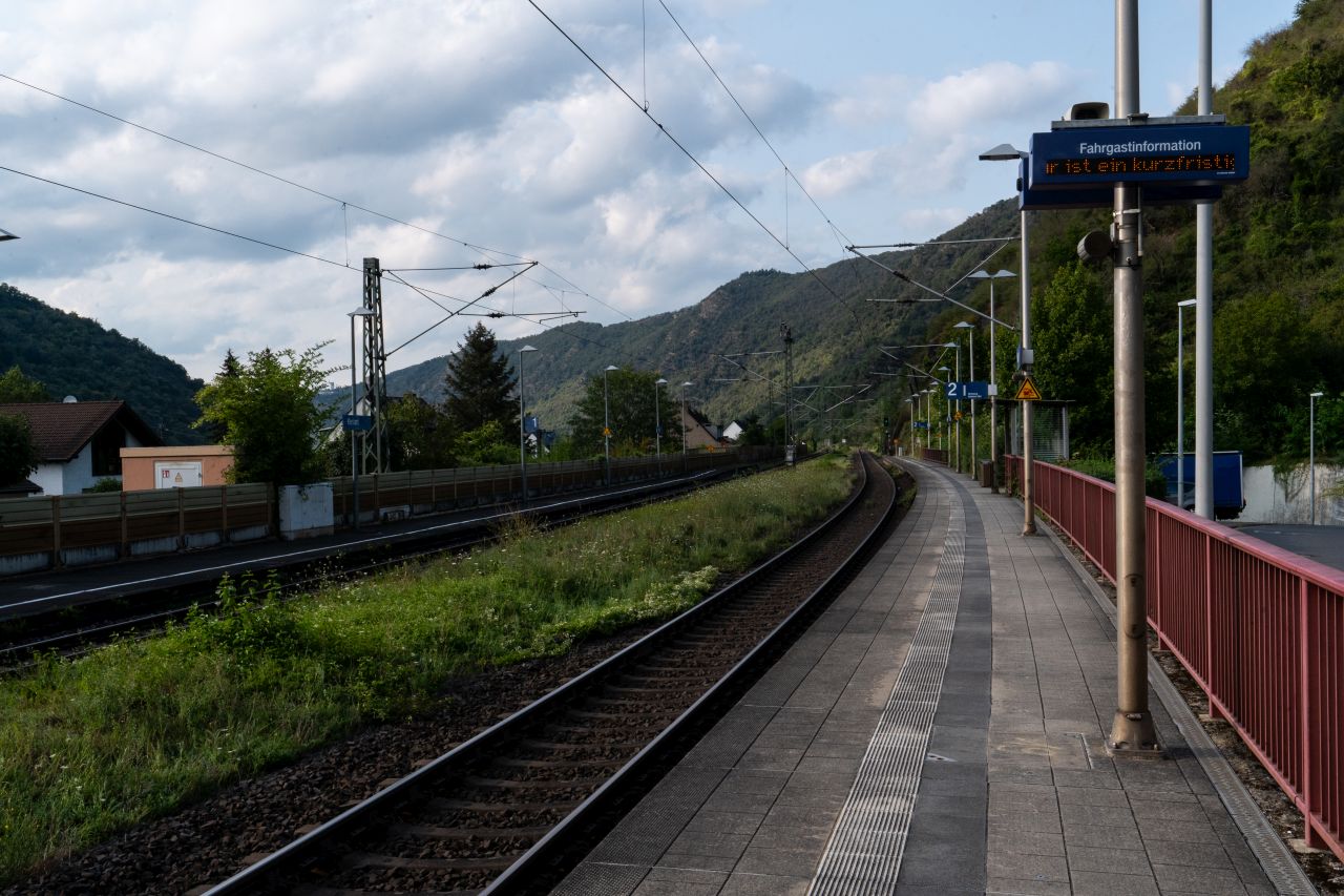 Bahnsteig der Station Kestert mit Gleisen, umgeben von grünen Hügeln und einem bewölkten Himmel.