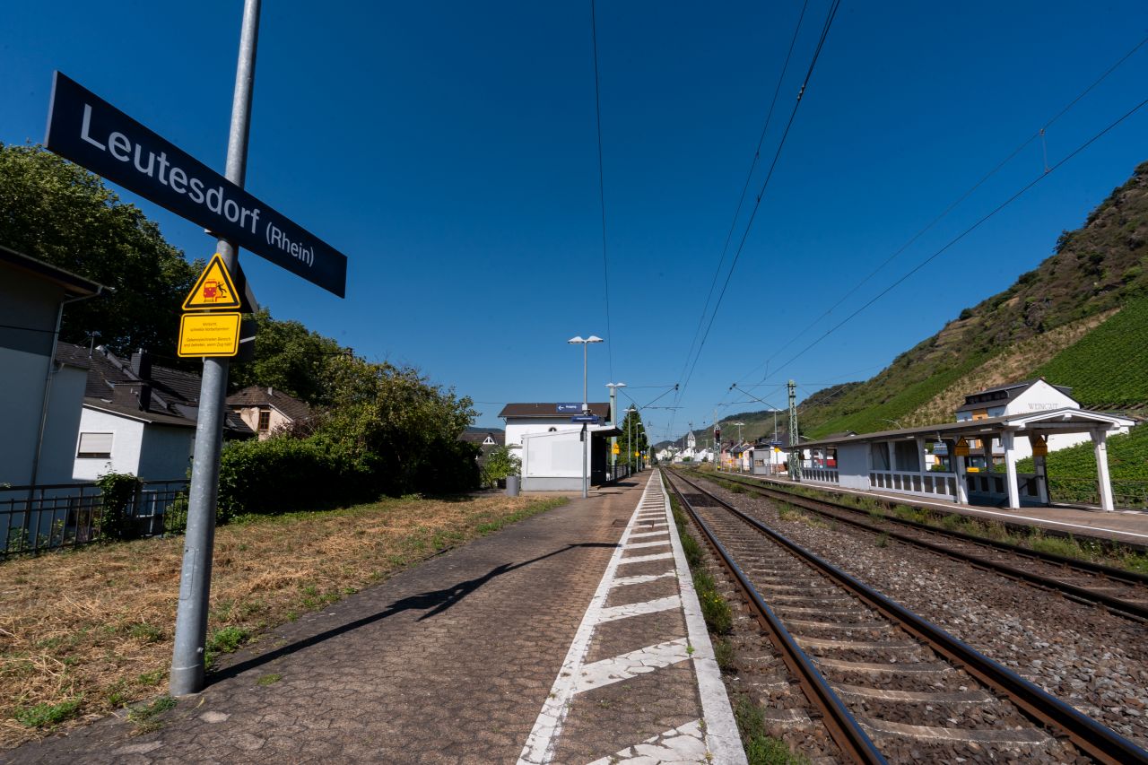 Ein Bahnsteig mit Gleisen und einem Schild "Leutesdorf (Rhein)", umgeben von Gebäuden und Weinbergen.