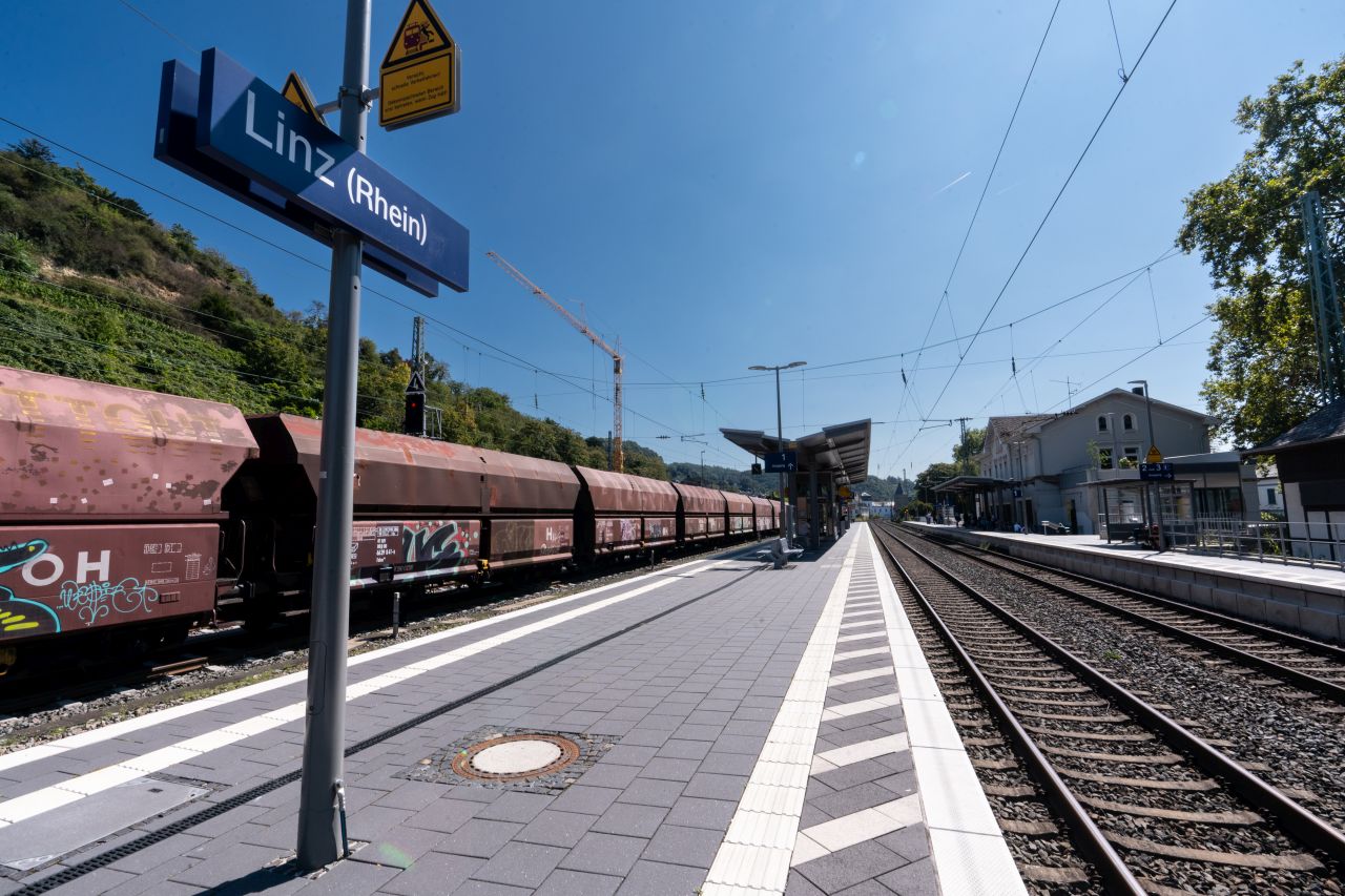 Bahnhof Linz (Rhein) mit zwei Gleisen, einem langsam passierenden Güterzug, einer Uhr und Sitzgelegenheiten auf dem Bahnsteig.