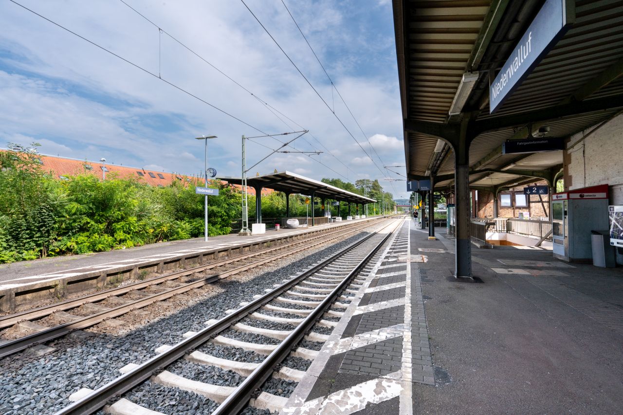 Bahnhof Niederwalluff mit zwei Gleisen und Bahnsteigen, einem Grünstreifen und angedeuteten Häusern im Hintergrund.