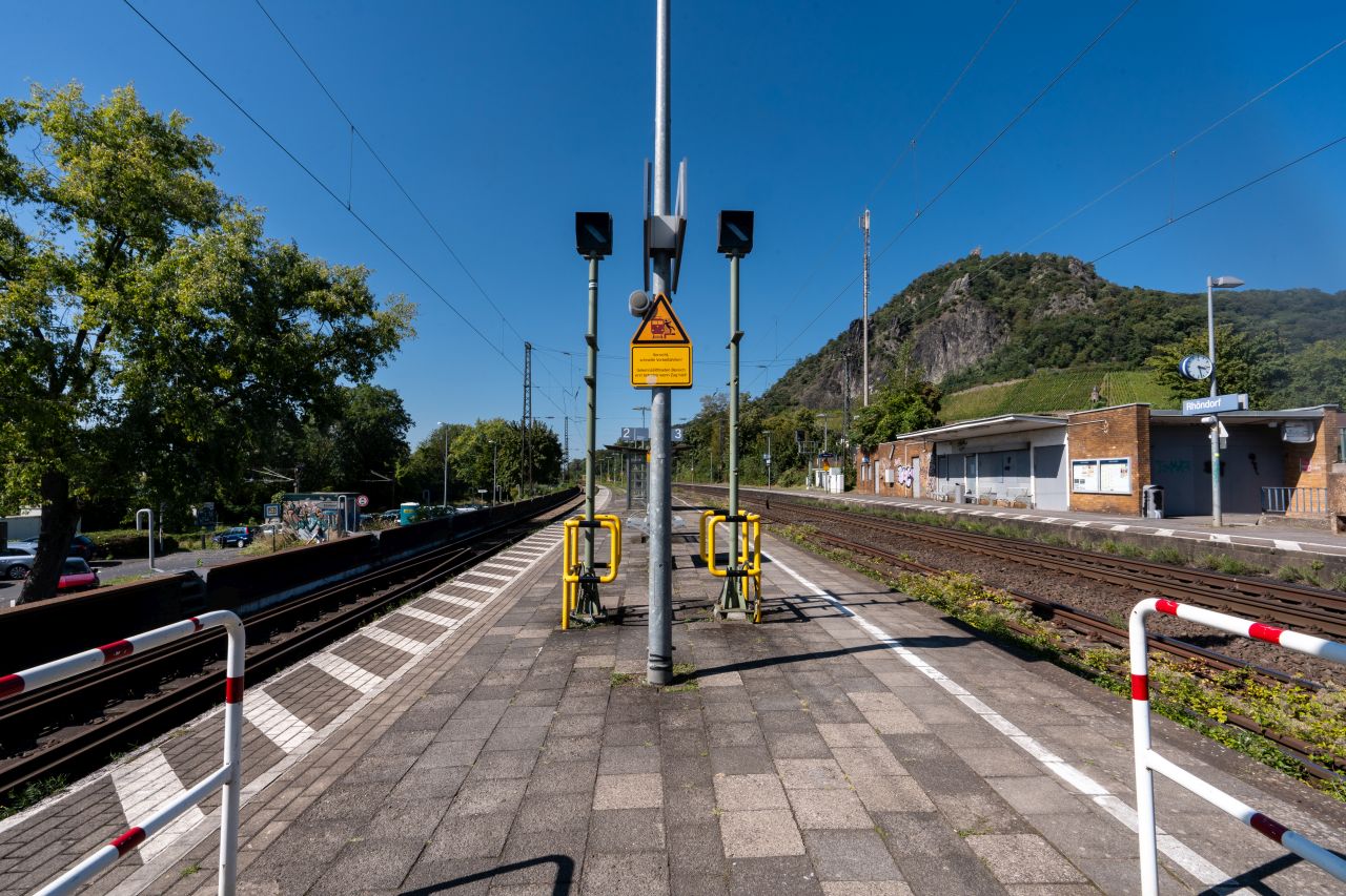Bahnsteig mit Gleisen auf beiden Seiten, Sicherheitsbarrieren und Warnschildern in der Mitte sowie einem grünen Hügel mit Weinbergen im Hintergrund.
