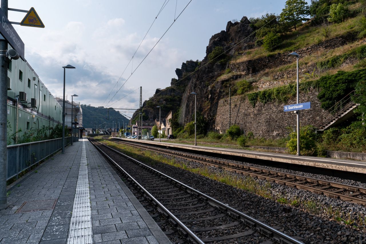 Bahnhof St. Goarshausen mit Bahngleisen und -Steigen mit Blick auf Hügel und eine felsige, begrünte Landschaft.