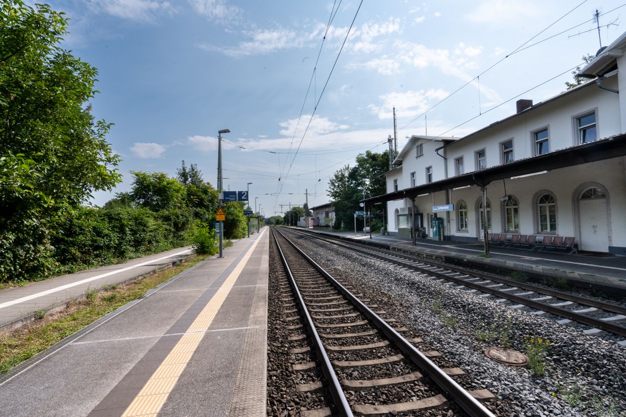 Leerer Bahnsteig des Bahnhof Wiesbaden Biebrich mit zwei Gleisen, einem weißen Bahnhofsgebäude und Bäumen im Hintergrund.